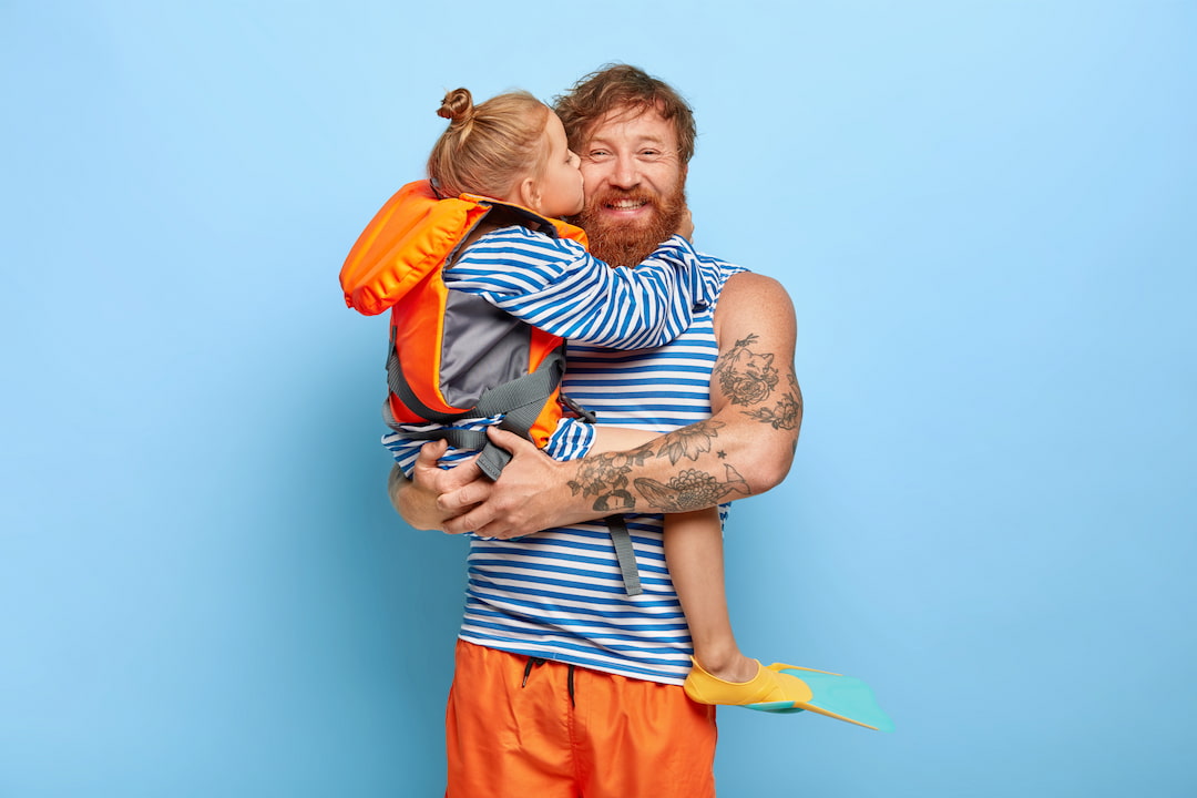 Mann im gestreiften Shirt hält Kind mit Schwimmweste und Flosse, Kind küsst ihn auf die Wange.“